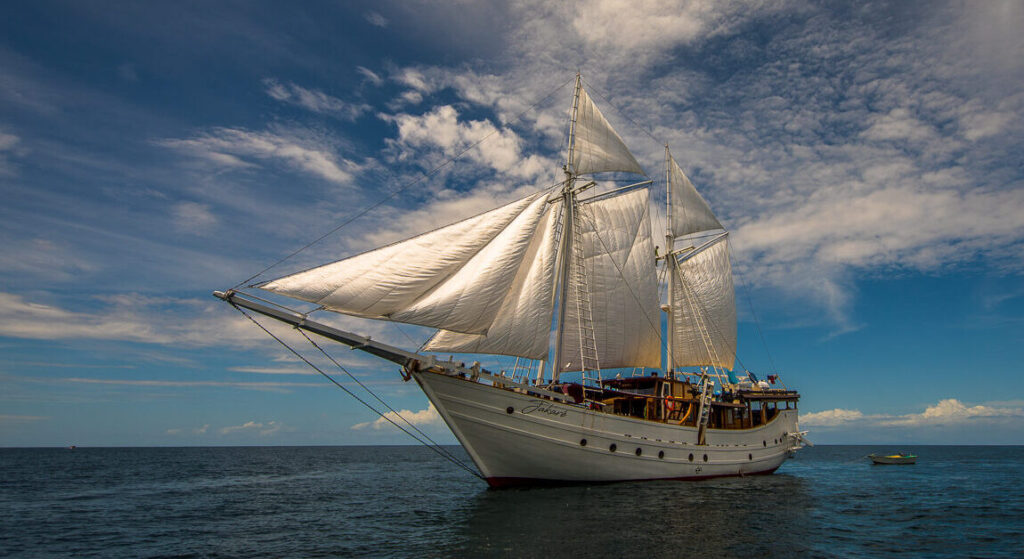 Side view of a traditional Indonesian phinisi boat, Jakare, with white sails fully open against a clear blue sky.